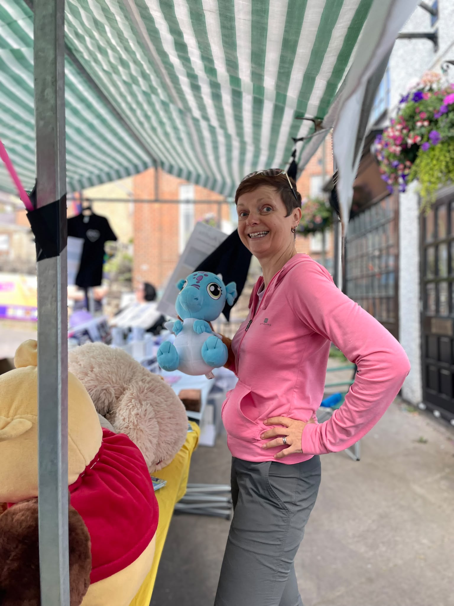 Volunteer in a pink hoodie holding a blue plush dragon toy under the market stall awning